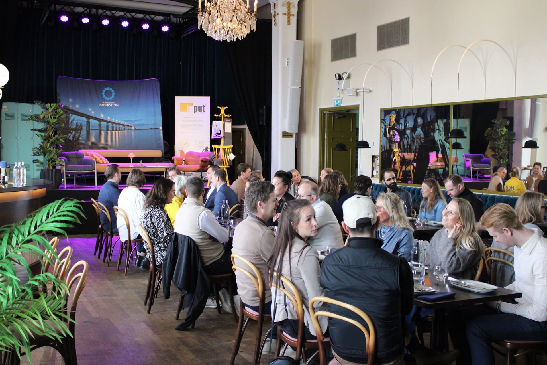 Gäster vid långbord i en sal med kristallkrona och scen under Promotionlunch